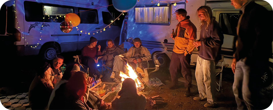 Friends gathered around a campfire between their vans at night